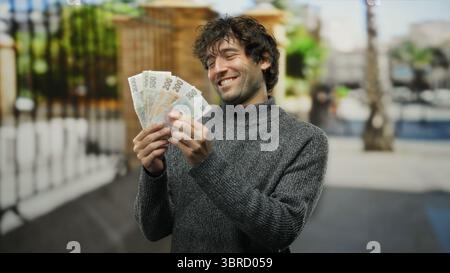 Ein lächelnder Mann, der auf einer Stadtstraße tschechische Kronen hält, zeigt sein spanisches Erbe und seinen urbanen Lebensstil im Freien. Stockfoto