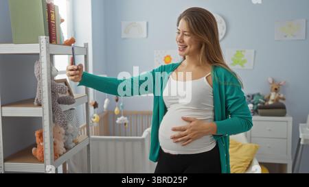 Schwangere Frau, die Selfie in einem gemütlichen Babyzimmer mit Spielzeugregal und Wiege macht, was einen glücklichen, erwartungsvollen Moment drinnen anzeigt. Stockfoto
