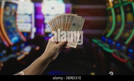 Ein Mann hält südkoreanische Won-Banknoten in einem Casino mit farbenfrohen Spielautomaten im Hintergrund, die den Nervenkitzel des Glücksspiels und des Geldes hervorheben Stockfoto