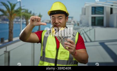 Junger Mann an der Strandpromenade mit Sicherheitsweste und Schutzhelm, der mit einer Hand die Daumen nach unten zeigt und uns in der anderen Dollars hält. Stockfoto