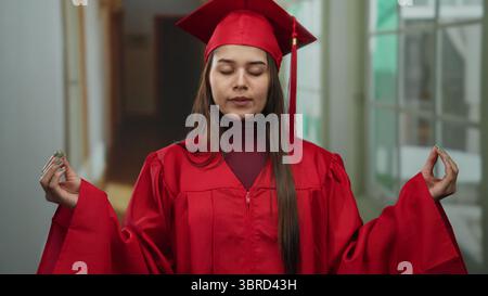 Junge Frau in rotem Abschlusskleid, die im Hotelflur meditiert, symbolisiert Ruhe und Leistung drinnen. Stockfoto