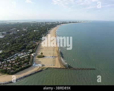 Blick aus der Vogelperspektive auf den goldenen Sand trifft auf das türkisfarbene Meer, gesäumt von hübschen Reihen von Sonnenliegen, neben den lebhaften grünen Bäumen, Lignano Sabbiadoro, Friaul-Julisch Venetien, Italien. Stockfoto