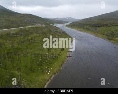 Aus der Vogelperspektive auf den dunklen, gewundenen Fluss, der durch das lebhafte grüne Tal fließt, flankiert von Hügeln unter gedämpftem Himmel, Karasjok, Finnmark, Norwegen. Stockfoto