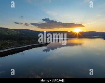 Aus der Vogelperspektive auf die goldenen Sonnenstrahlen, die den ruhigen See küssen und sich wie flüssiges Gold vom Wasser spiegeln, während der Wald in tiefen smaragdgrünen Tönen wacht, Schluchsee, Baden-Württemberg, Deutschland. Stockfoto