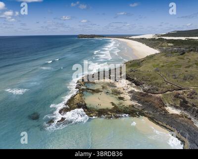 Aus der Vogelperspektive treffen türkisfarbenes Wasser auf zerklüftete, dunkle Felsen und einen unberührten Sandstrand unter hellem Himmel, Queensland, Australien. Stockfoto