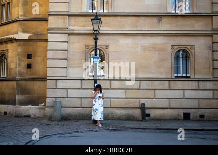 Eine Frau in einem Blumenkleid steht an einem Lampenpfosten vor einem historischen Sandsteingebäude in Cambridge, Großbritannien, die in ihr Handy eingesogen ist Stockfoto