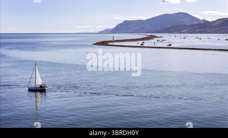 Aus der Vogelperspektive auf ein einsames Segelboot, das über das ruhige Wasser gleitet, wobei der ferne Hafen und die Berge einen ruhigen Kontrast aus kühlem Blau und sanftem Grün bilden, Nelson Region, Neuseeland. Stockfoto
