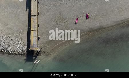 Aus der Vogelperspektive sehen Sie zwei pulsierende pinkfarbene Kajaks an einem grauen, steinigen Strand neben einem verwitterten Pier, wo das türkisfarbene Meer auf die Küste trifft, Nelson Region, Neuseeland. Stockfoto
