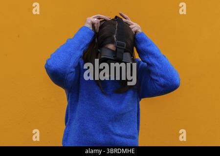Eine junge Frau in einem Jugendoutfit benutzt eine Virtual-Reality-Brille. Eine Brünette in Blau auf gelbem Hintergrund bringt ihr ein vr-Gadget auf den Kopf. Eintauchen in ein VI Stockfoto