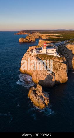 Aus der Vogelperspektive auf den zerklüfteten, sonnenverwöhnten Klippen vor dem tiefblauen Ozean, einem Leuchtfeuer am Horizont, Sagres, Faro, Portugal. Stockfoto