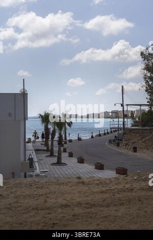 Blick auf das türkisfarbene Wasser trifft auf die Sandküste, Palmen säumen die Straße in einer Küstenszene, ein ruhiger Moment gefrorener Zeit, Varosha, Famagusta, C Stockfoto
