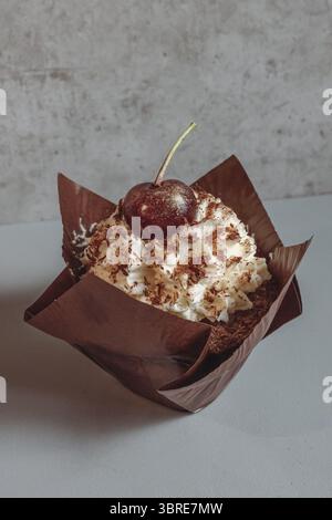 Schokoladenmuffin mit Kirschen und Sahne in einer braunen Verpackung. Süße Versuchung. Restauranthintergrund. Essen in einem Café. Dessert mit Kaffee. Stockfoto