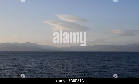 Meereswellen schlagen sanft gegen die Küste, während Wolken in der Dämmerung über ferne Berge gleiten. Sanfte Farben prägen den Himmel und schaffen eine friedliche Atmosphäre am Abend. Stockfoto