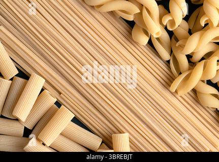 Aus nächster Nähe sehen Sie verschiedene ungekochte Nudelsorten wie Spaghetti, Fusilli und Rigatoni, die auf einer dunklen Oberfläche angeordnet sind Stockfoto