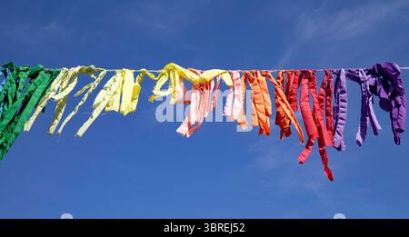 Regenbogenfarbene Lumpen oder Wimpel, auf einer Linie aufgereiht, fliegen im Wind gegen einen blauen Himmel Stockfoto