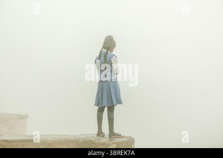 New Delhi, Indien - 24. Juni 2025: Blick auf ein einsames Mädchen in Schuluniform, das auf einem Felsvorsprung steht und in einen dichten, ätherischen Nebel blickt. Stockfoto