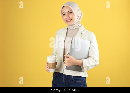 Lächelnde junge asiatische Hijab-Frau in Creme-Anzug und Hijab mit Laptop und heißem Kaffee. Isoliert auf gelbem Hintergrund Stockfoto