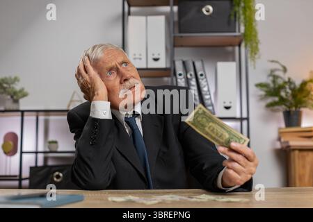 Kaukasischer Senior-Geschäftsmann Freelancer zählt Bargeld zu wenig Geld. Finanzkrise. Konkurs. Armut und Elend. Großvater im Anzug sitzt am Tisch im Büro. Stockfoto