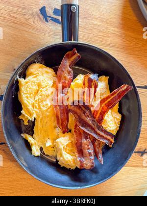 Gekochte Speckstreifen ruhen auf perfekt gerührten Eiern in einer schwarzen Pfanne. Das appetitliche Frühstück wird auf einem Holztisch serviert, der einen betont Stockfoto