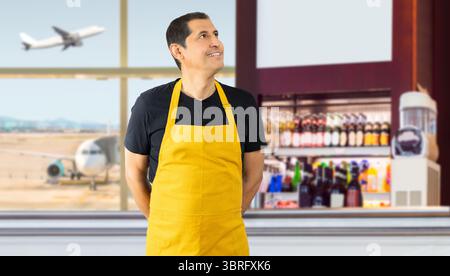 Hispanischer Barista mit gelber Schürze schaut auf den Kopierraum, den Mitarbeiter des Lieferdienstes, den Kellner in gelber Schürze im Café eines Flughafens Stockfoto