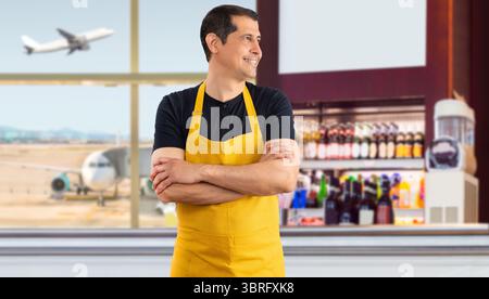 Hispanischer Barista steht lächelnd zur Seite und blickt mit gelber Schürze in den Coffee Shop eines Flughafens Stockfoto