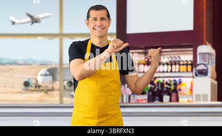 Hispanischer Barista mit gelber Schürze stehend und lächelnd und blickt auf die Kamera und zeigt mit zwei Händen und Fingern zur Seite im Café A Stockfoto