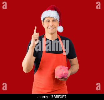 Studio-Aufnahme eines Mannes mit roter Schürze eines Angestellten, der eine Schweinebank vor rotem Hintergrund hält, während er einen Weihnachtshut trägt. Stockfoto
