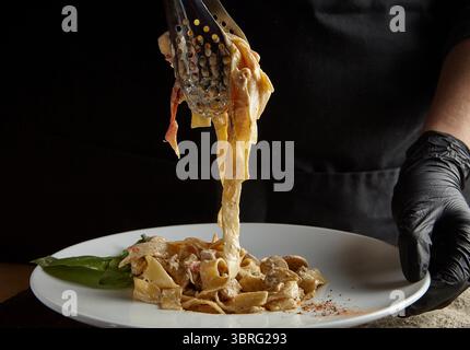 Koch serviert cremige Pasta auf einem weißen Teller mit einer Küchenzange und trägt schwarze Handschuhe Stockfoto