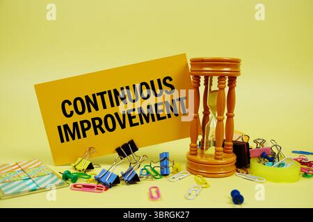 Continuous Improvement Text auf Papierkarte mit Sanduhr und Briefpapier auf gelbem Hintergrund Stockfoto