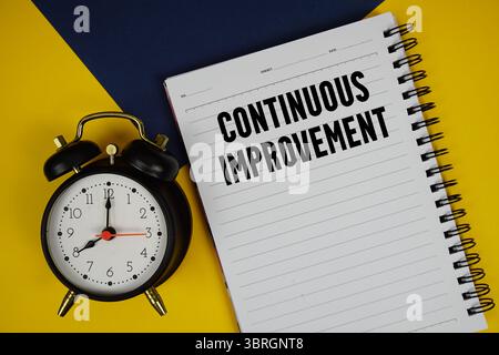 Text zur kontinuierlichen Verbesserung auf Notizblock mit Wecker auf gelbem Hintergrund Stockfoto