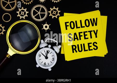 Befolgen Sie die Textnachricht „Sicherheitsregeln“ auf Haftnotiz mit Vergrößerungsglas und Wecker auf schwarzem Hintergrund Stockfoto