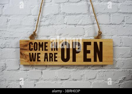 Kommen Sie herein Wir sind offen Text auf hölzernem Schild, das auf weißem Backsteinmauerhintergrund hängt Stockfoto