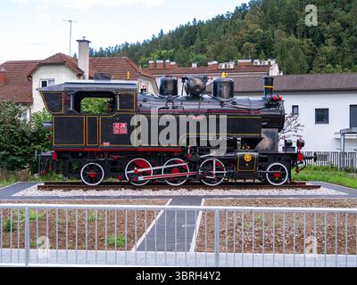 Eine alte schwarze Dampflokomotive, die draußen auf einem Gleisabschnitt ausgestellt wurde, umgeben von einem kleinen Zaun, mit Wohngebäuden und Bäumen im Hinterbau Stockfoto