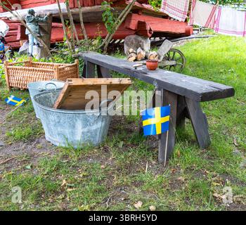Sodertalje, Schweden - 6. Juni 2025: Eine rustikale Outdoor-Szene mit einer Holzbank, Metalleimern und schwedischen Fahnen. Die Umgebung umfasst Grünanlagen und Stockfoto