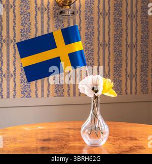 Eine schwedische Flagge steht aufrecht in einer klaren Glasvase, gefüllt mit bunten Blumen auf einem Holztisch. Der Hintergrund ist mit einer gemusterten Tapete, cre, versehen Stockfoto