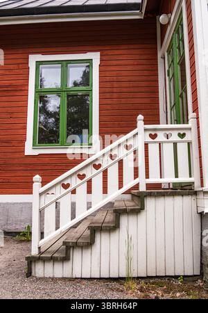 Sodertalje, Schweden - 6. Juni 2025: Ein charmantes Holzhaus mit roter Fassade und grünen Fenstern. Die weiße Treppe ist mit herzförmigen Ausschnitten versehen Stockfoto