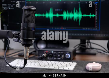 Nahaufnahme des schwarzen Kondensatormikrofons und der Audioschnittstelle auf dem Schreibtisch mit grünen Klangformen auf dem Monitor, die modernes Podcasting und Musik hervorheben Stockfoto