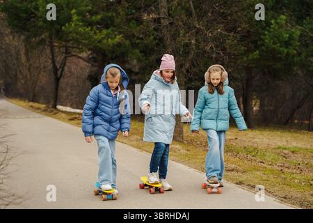Teenagerinnen fahren Skateboards auf dem Stadtweg im Herbst-Winterpark. Jacken und Jeans spiegeln den urbanen Jugendstil wider. Straßensport verbessert Balance, Bewegung, Stockfoto