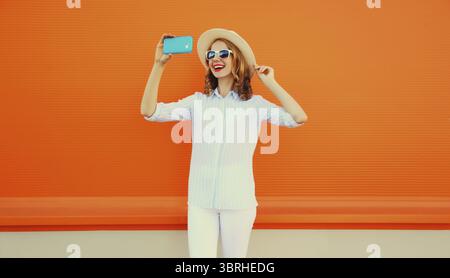 Porträt einer glücklichen lächelnden jungen Frau, die Selfie mit Smartphone macht und Strohhut auf orangem Hintergrund trägt Stockfoto
