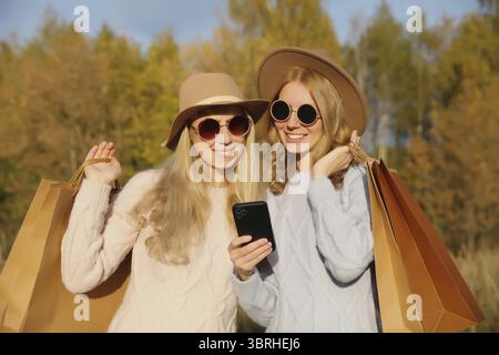 Modische glückliche junge zwei Frauen, die mit Einkaufstaschen, Smartphone halten, stylische bunt lächelnde Freundinnen, die das Telefon zusammen halten o Stockfoto