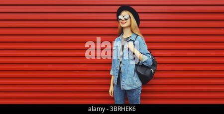 Porträt einer glücklichen lächelnden jungen Frau, die wegschaut mit schwarzem Rundhut, jeansjacke, Rucksack auf rotem Hintergrund, leerer Kopierraum für Werbung Stockfoto