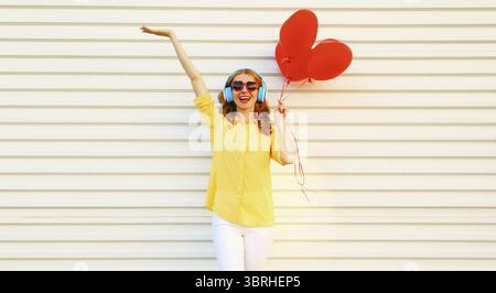 Porträt einer glücklichen lächelnden jungen Frau, die Musik in Kopfhörern hört, die einen Haufen roter herzförmiger Ballons auf weißem Hintergrund hält Stockfoto