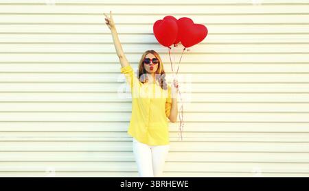 Porträt der schönen jungen Frau mit rotem herzförmigem Ballon, der ihre Lippen bläst und einen süßen Luftkuss sendet, der Sonnenbrille auf weißem Hintergrund trägt Stockfoto