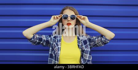 Porträt einer modernen stilvollen jungen Frau in weißer Brille, lässige Kleidung, die auf blauem Wandhintergrund posiert Stockfoto