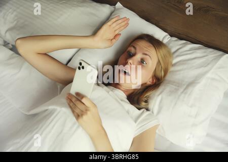 Überraschte junge Frau mit Telefon, das zu Hause im Bett auf Kissen lag Stockfoto