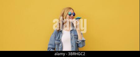 Glückliche junge Frau, die am Telefon spricht, Sprachaufzeichnungsgerät benutzt, Assistenten benutzt oder Anrufe auf dem Smartphone in lässiger Kleidung auf dem hellen YE entgegennimmt Stockfoto