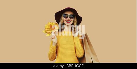 Herbstfarben-Outfit, Porträt einer schönen jungen Frau mit Einkaufstasche hält gelbe Ahornblätter tragen Hut, Strickpullover auf beigem Studio Stockfoto