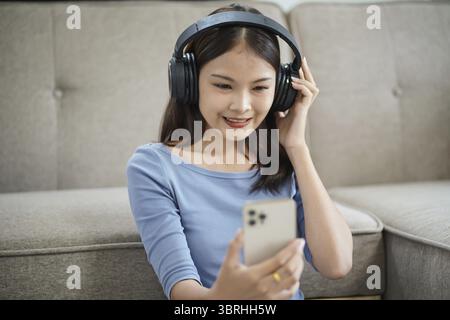 Entspannte junge schöne asiatische Frau Musik-Liebhaber hört Musik auf Ohrhörern hält Handy im Wohnzimmer hört Musik mit Kopfhörern und m Stockfoto