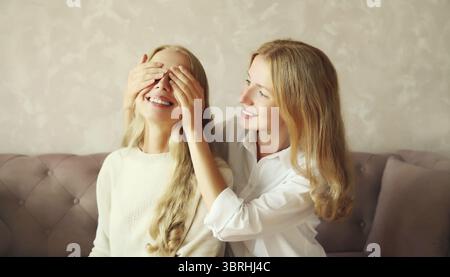 Glückliche lächelnde weiße erwachsene Tochter, die die Augen der Mutter bedeckt, überraschend auf der Couch zu Hause sitzend Stockfoto