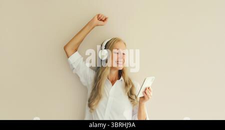 Glückliche, fröhliche Frau mittleren Alters, die Musik mit Mobiltelefon in den Kopfhörern hört und Spaß beim Tanzen auf Studiohintergrund hat Stockfoto